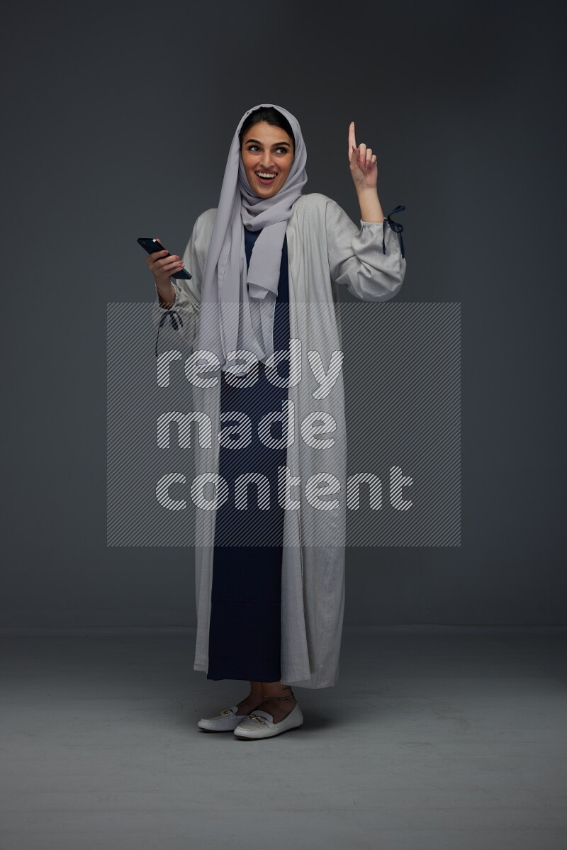 A Saudi woman wearing a light gray Abaya and head scarf standing and holding a phone on a grey background