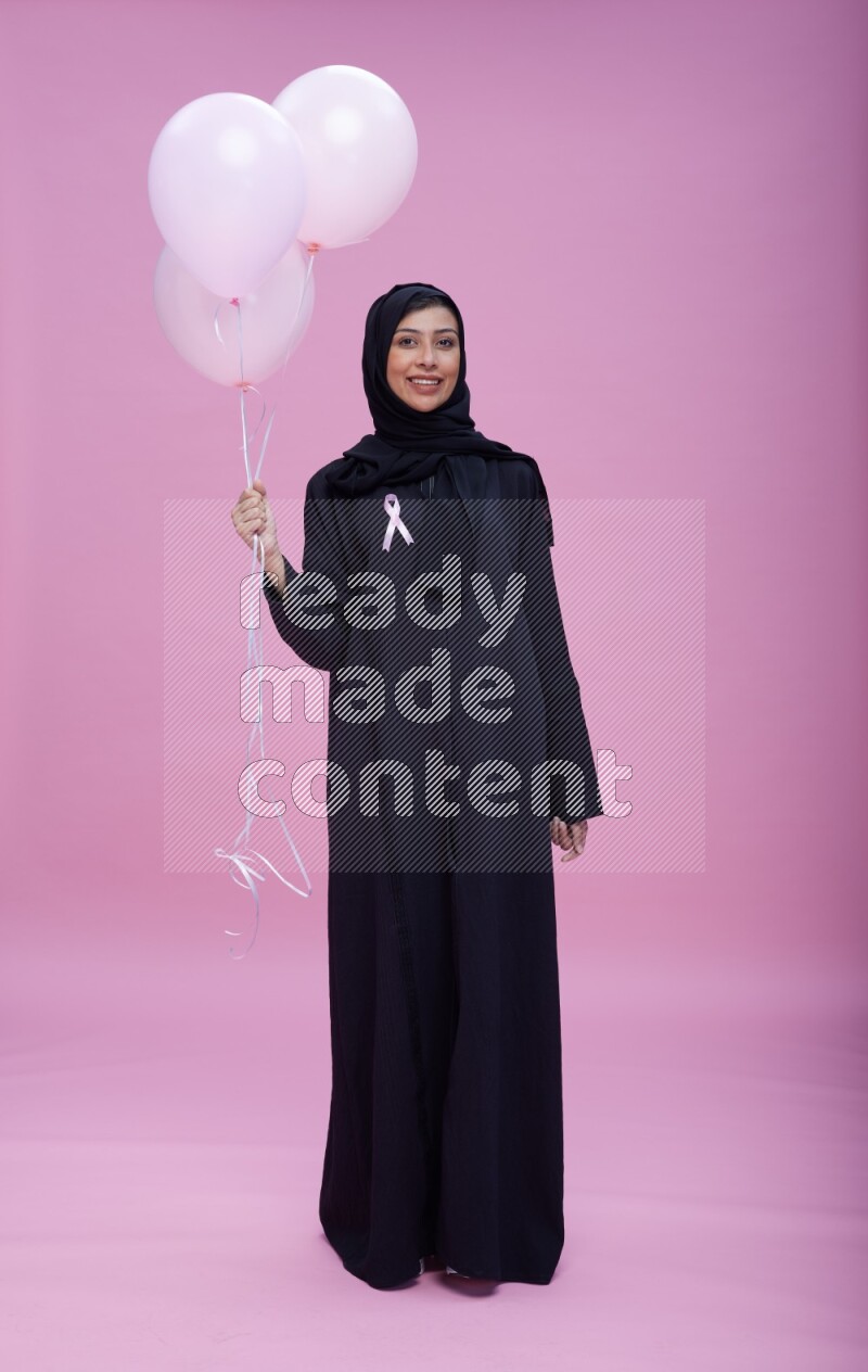 Saudi woman wearing pink ribbon on Abaya standing holding balloons on plain pink background