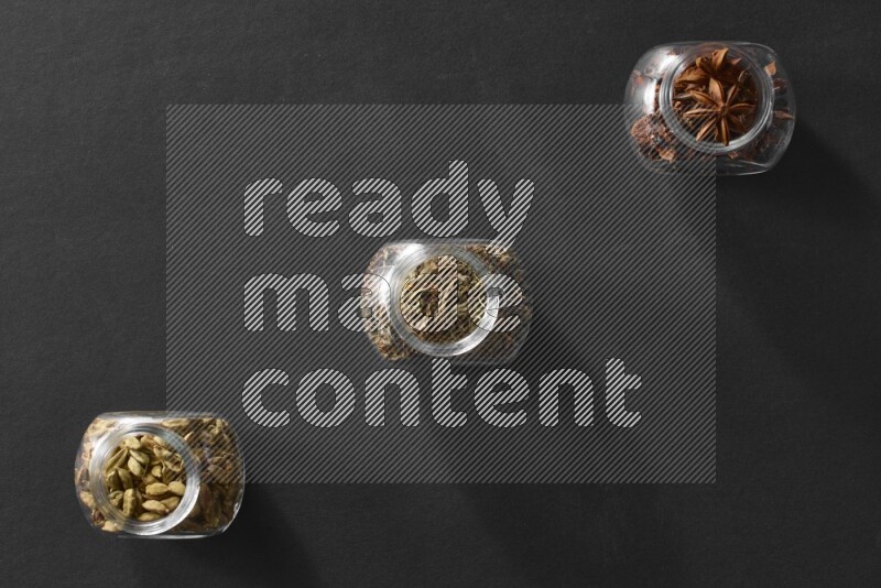 3 glass spice jars full of star anise, basil and cardamom on black flooring