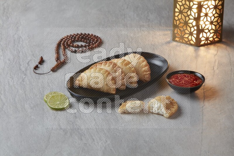 Four fried sambosas in an oval shaped black plate, beside a cut cheese sambosa, a brown misbaha and a golden lantern on a gray background