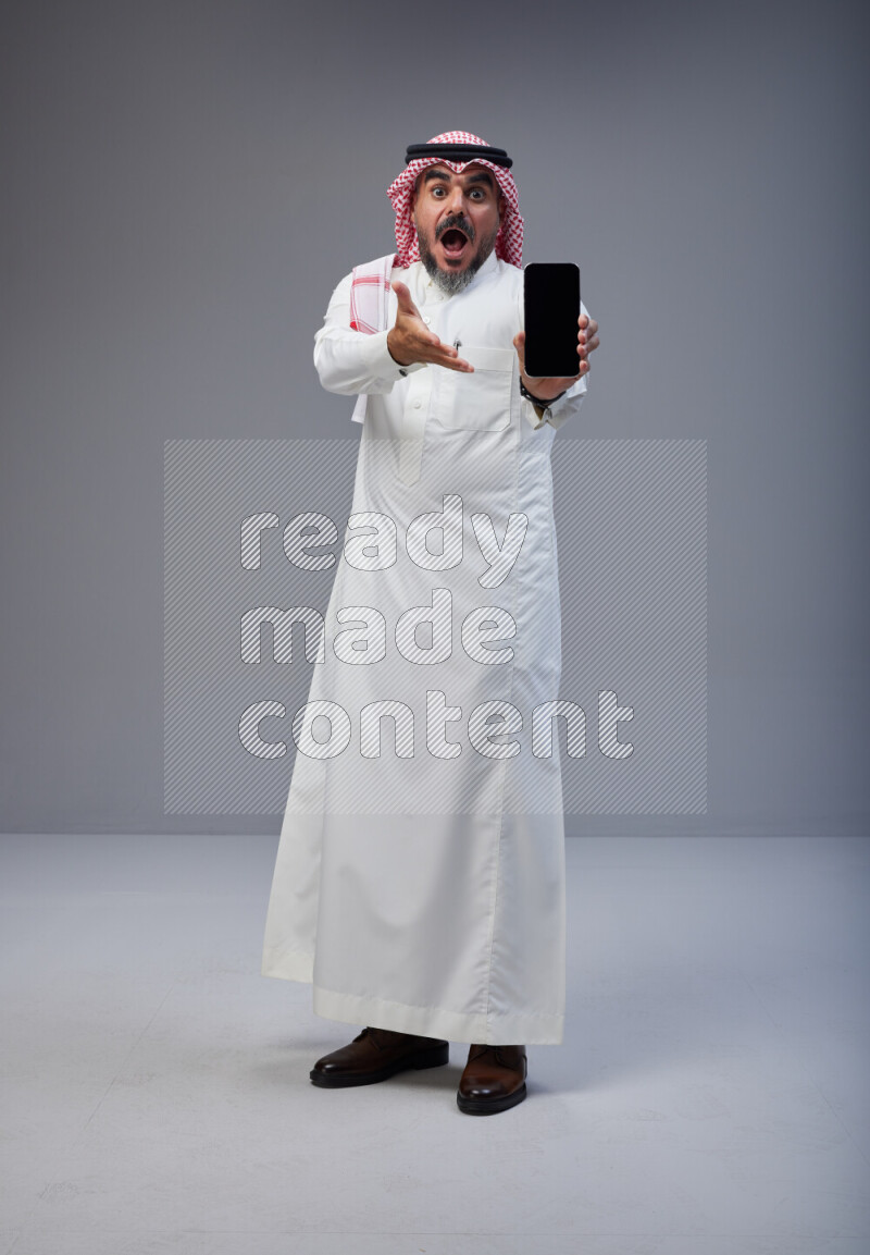 Saudi man Wearing Thob and red Shomag standing showing phone to camera on Gray background