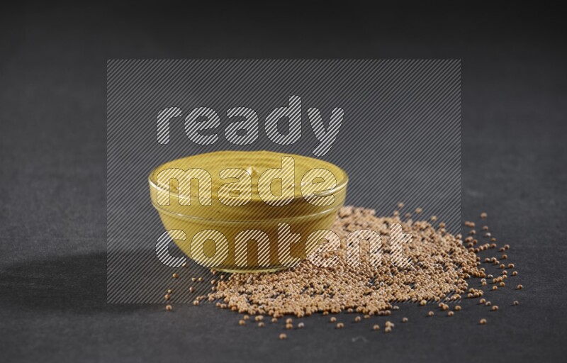 A glass bowl full of mustard paste with mustard seeds underneath on black flooring