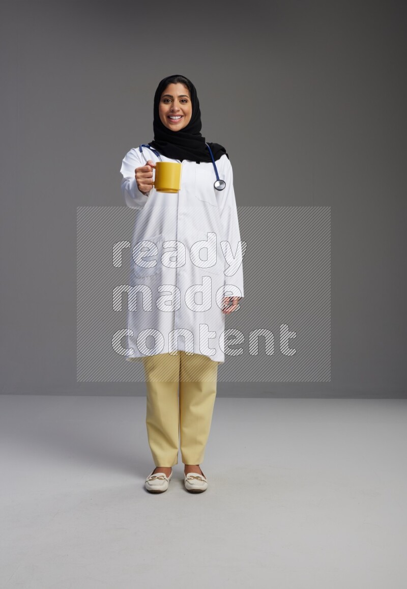 Saudi woman wearing lab coat with stethoscope standing holding mug on Gray background