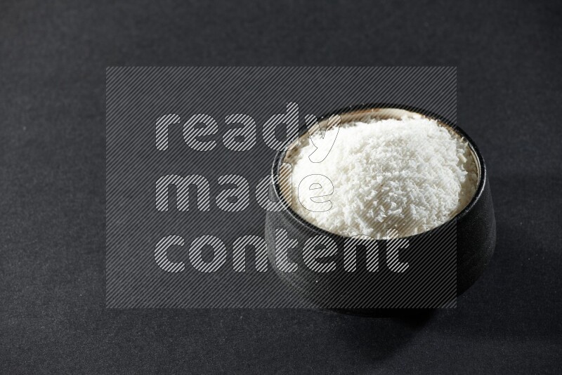 A black pottery bowl full of desiccated coconut on a black background in different angles