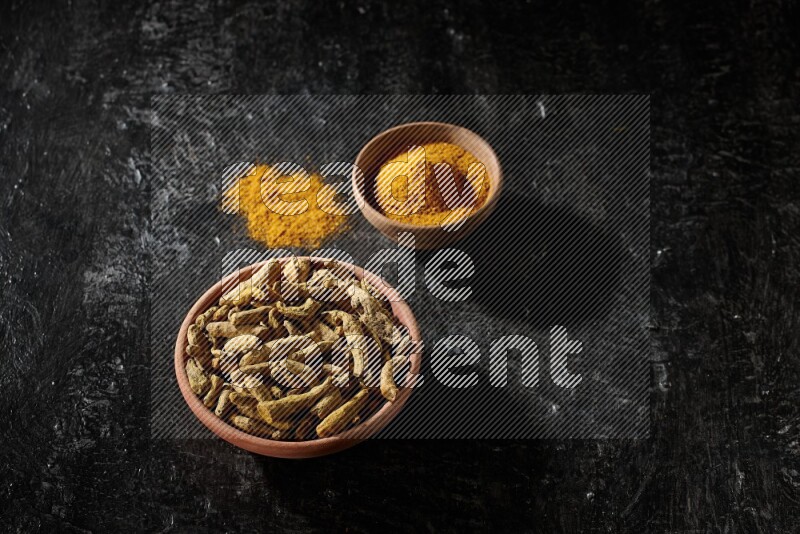 2 wooden bowls, one full of turmeric powder and the other full of dried turmeric whole fingers on a textured black flooring