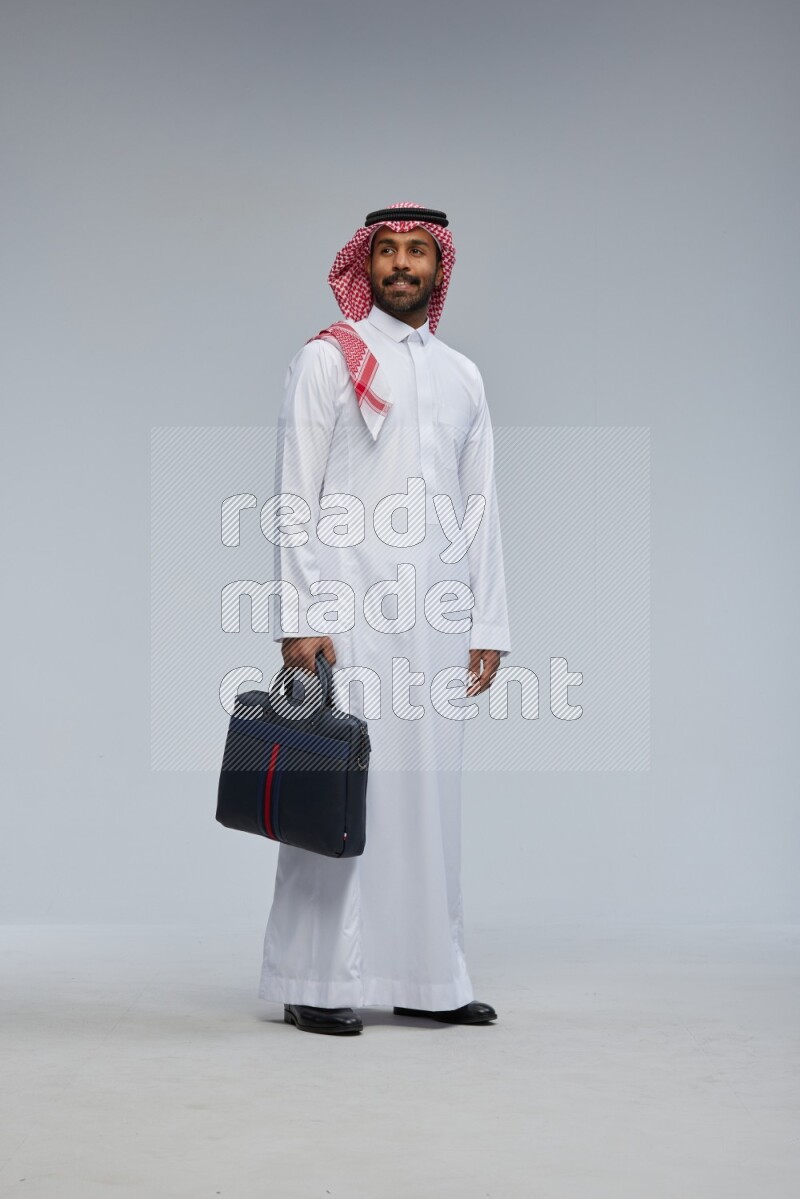 Saudi man Wearing Thob and shomag standing holding bag on Gray background