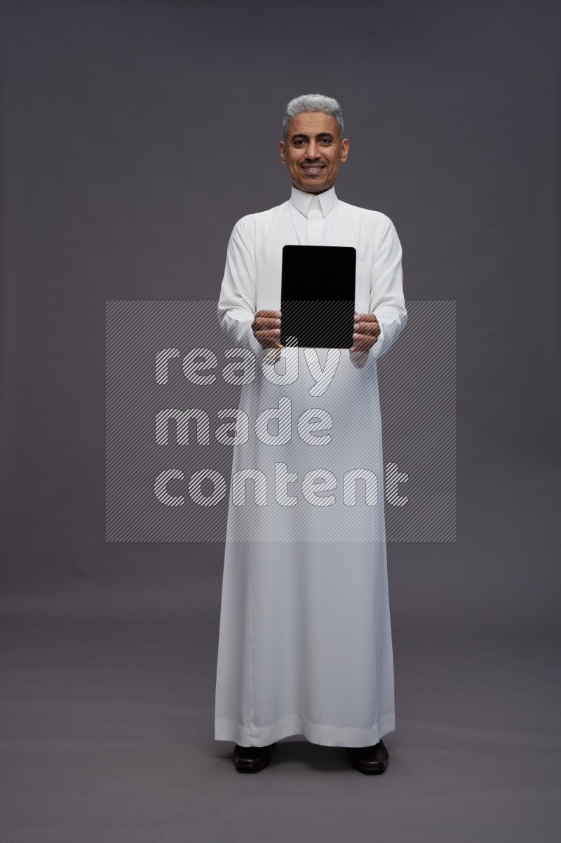 Saudi man wearing thob with neck strap employee badge standing showing tablet to camera on gray background