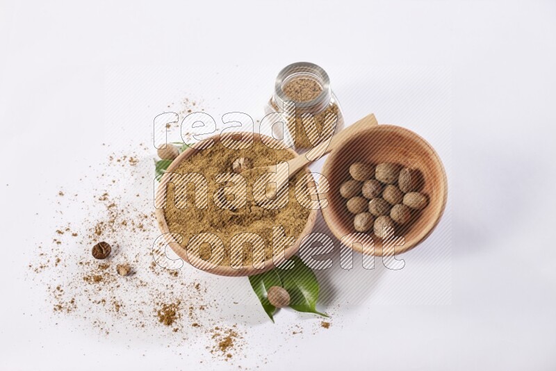 A wooden bowl with a spoon in it and glass spice jar all, full of nutmeg powder and a wooden bowl full of whole nutmeg seeds on a white flooring