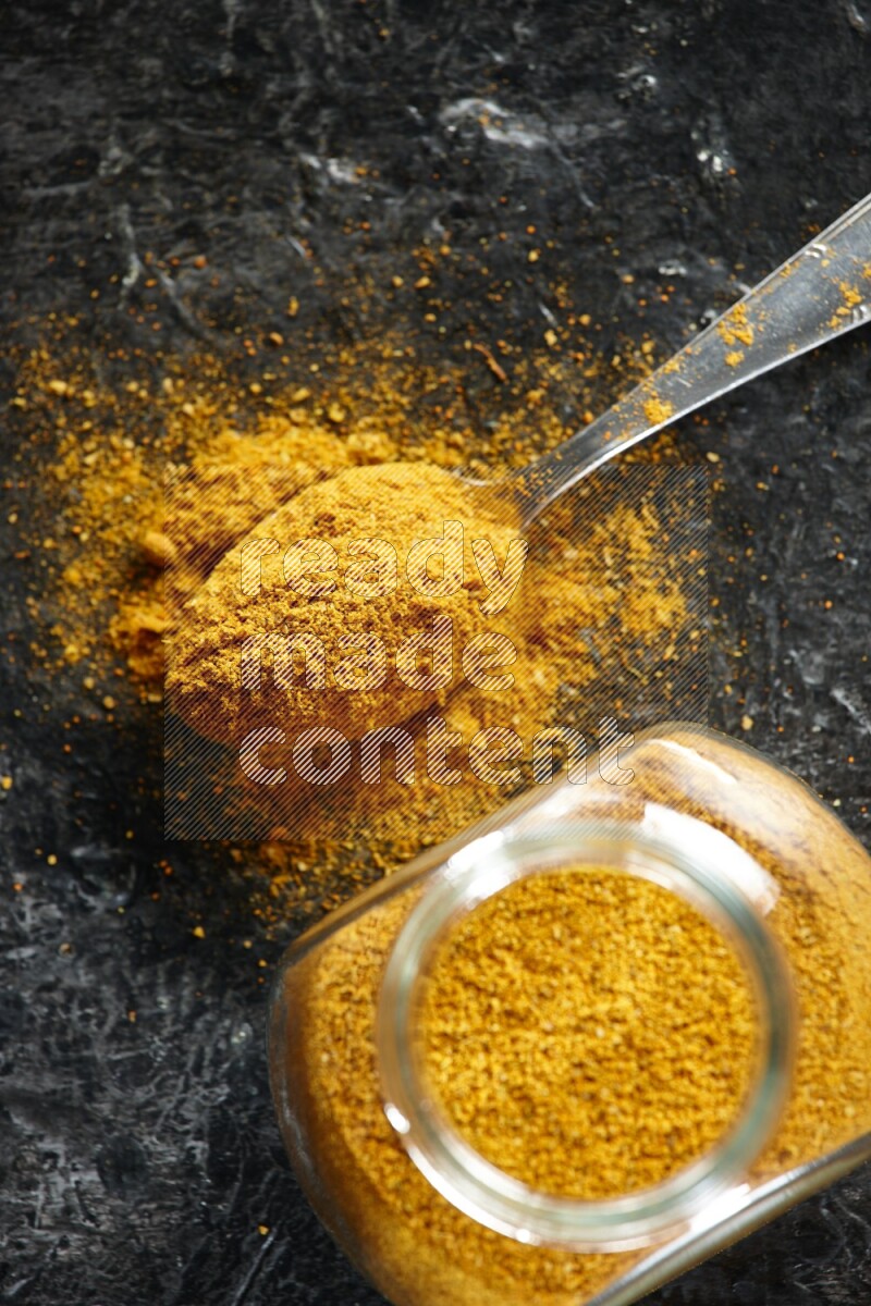 A glass spice jar and metal spoon full of turmeric powder on textured black flooring