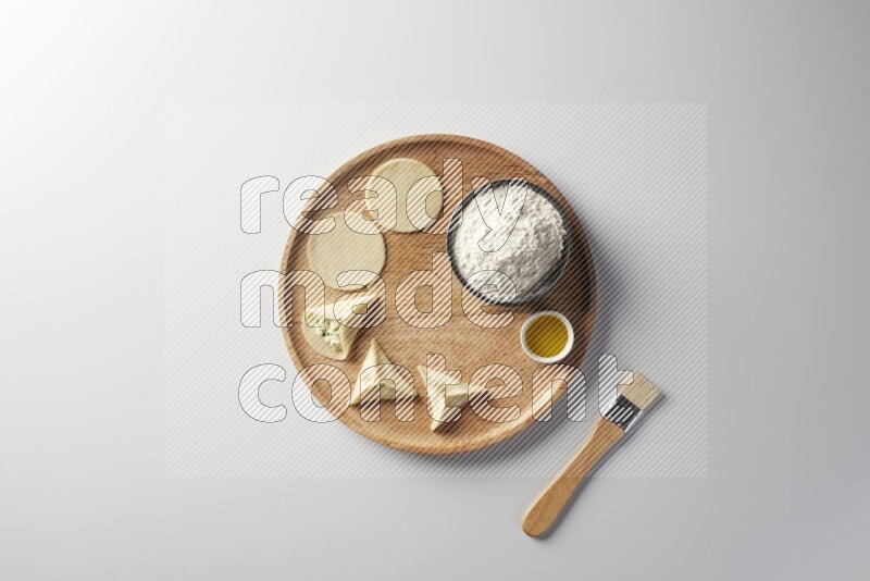 two closed sambosas and one open sambosa filled with cheese while flour, and oil with oil brush aside in a wooden dish on a white background