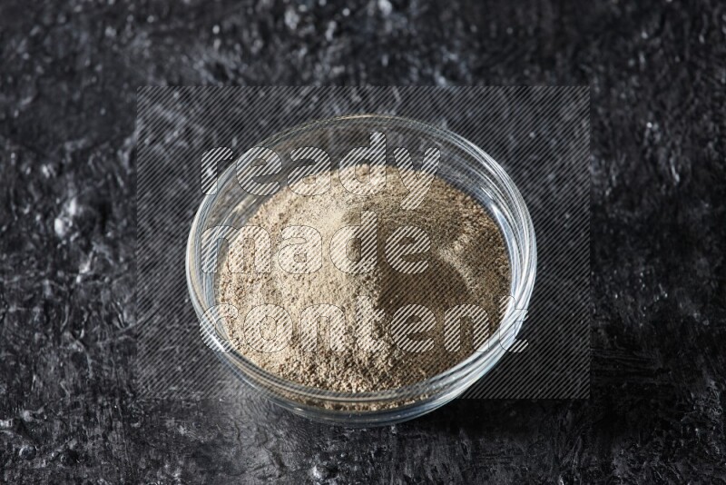 A glass bowl full of white pepper powder on textured black flooring