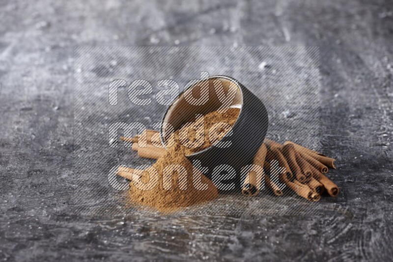 Black pottery bowl over filled with cinnamon powder and cinnamon sticks around the bowl on a textured black background