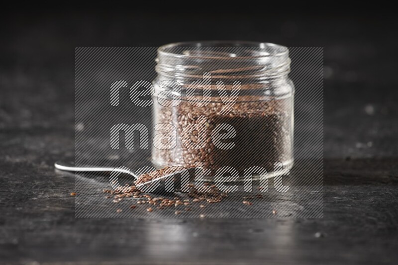 A glass jar full of flaxseeds with a metal spoon full of the seeds on a textured black flooring