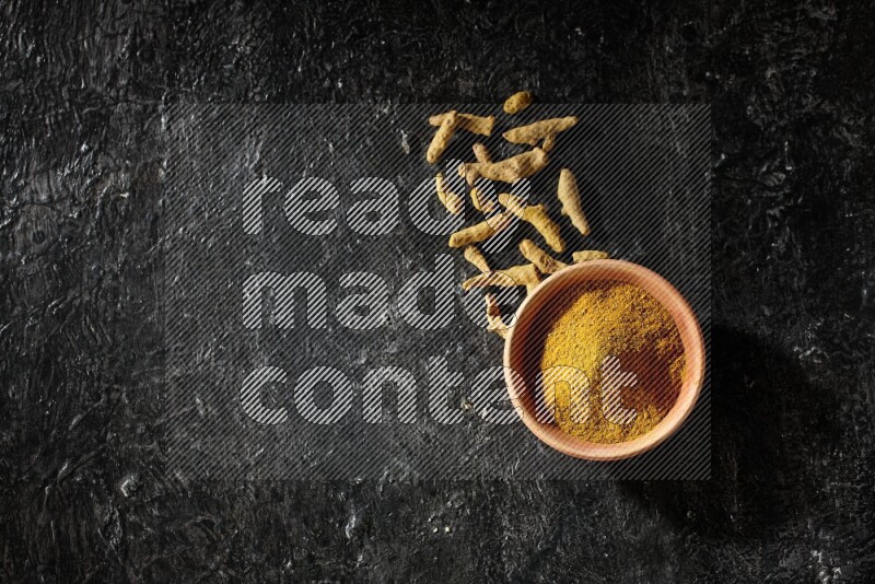 A wooden bowl full of turmeric powder with dried turmeric fingers on textured black flooring