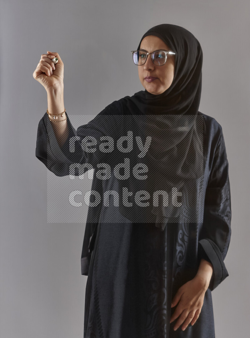 Woman in black abaya, hijab and glasses holding a marker pen to write on a transparent board with different reactions and expressions on grey background
