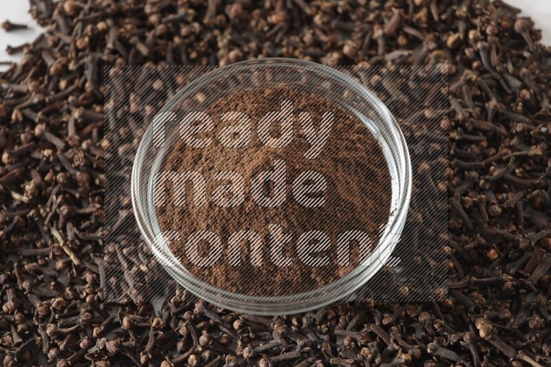 وعاء زجاجي ممتلئ ببودرة القرنفل وحبوب القرنفل متناثرة على أرضية بيضاء