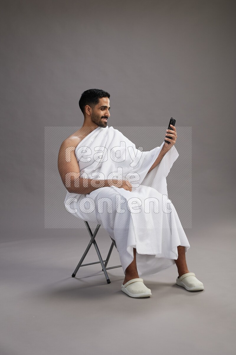 A man wearing Ehram sitting on chair taking selfie on gray background