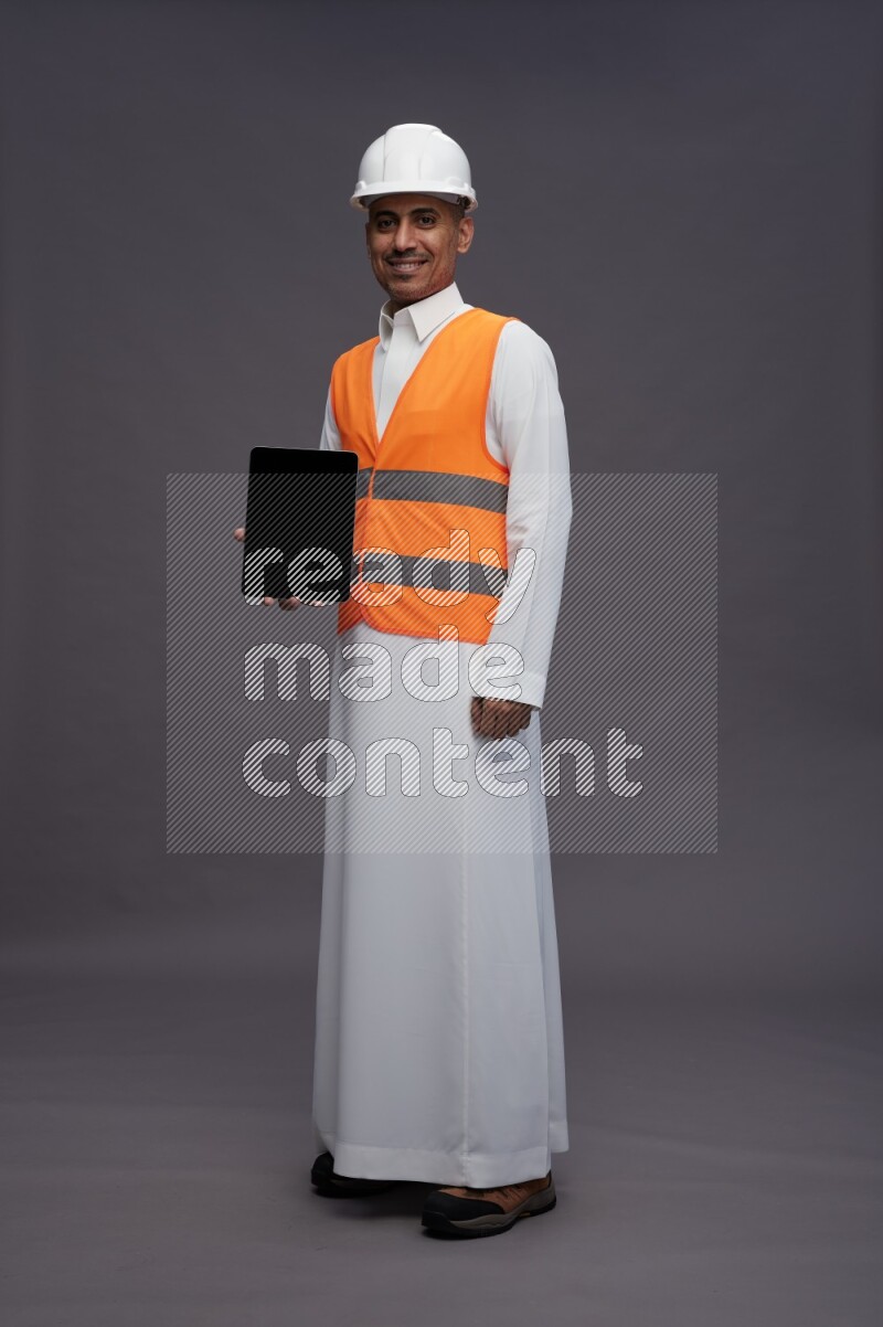 Saudi man wearing thob with engineer vest standing showing tablet to camera on gray background