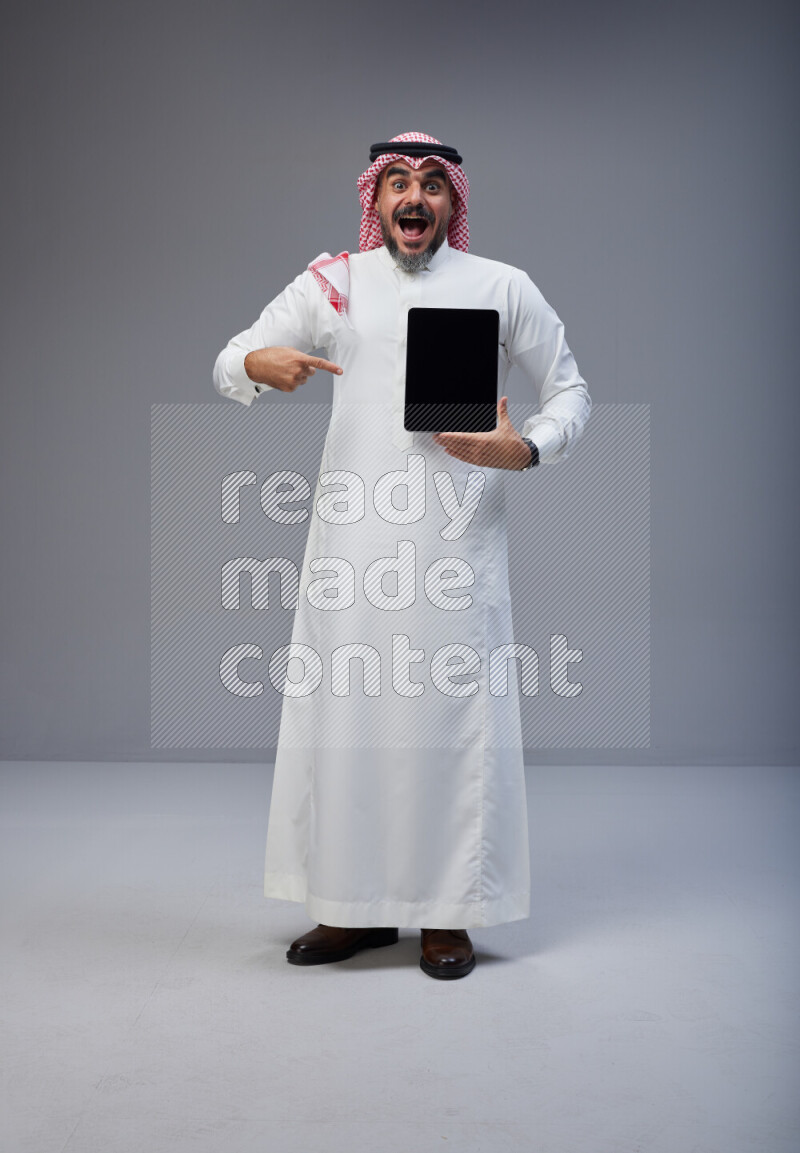 Saudi man Wearing Thob and red Shomag standing showing tablet to camera on Gray background