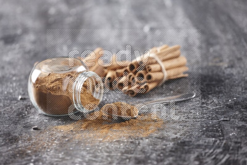 Herbal glass jar full cinnamon powder flipped and a metal spoon full of powder, cinnamon sticks stacked and bounded in the back on textured black background in different angles