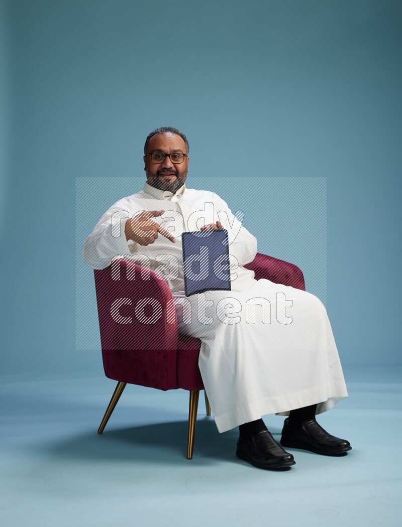 Saudi Man without shimag sitting on chair working on tablet on blue background