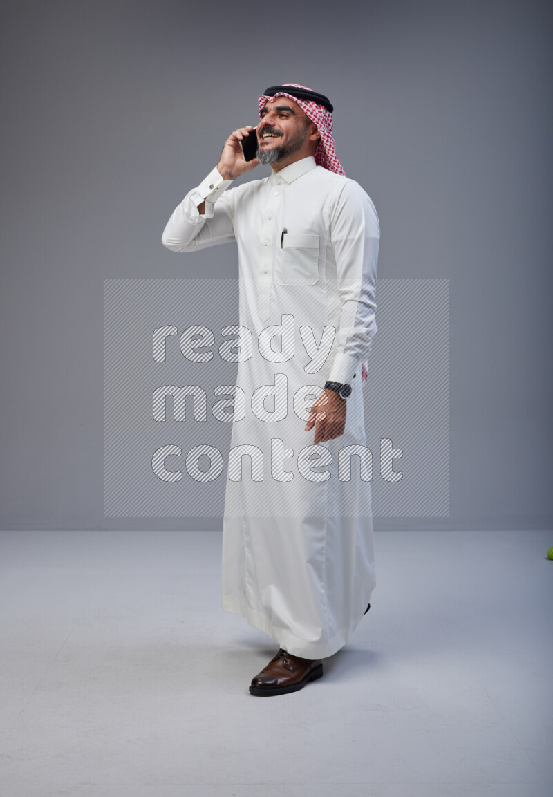 Saudi man Wearing Thob and red Shomag standing talking on phone on Gray background