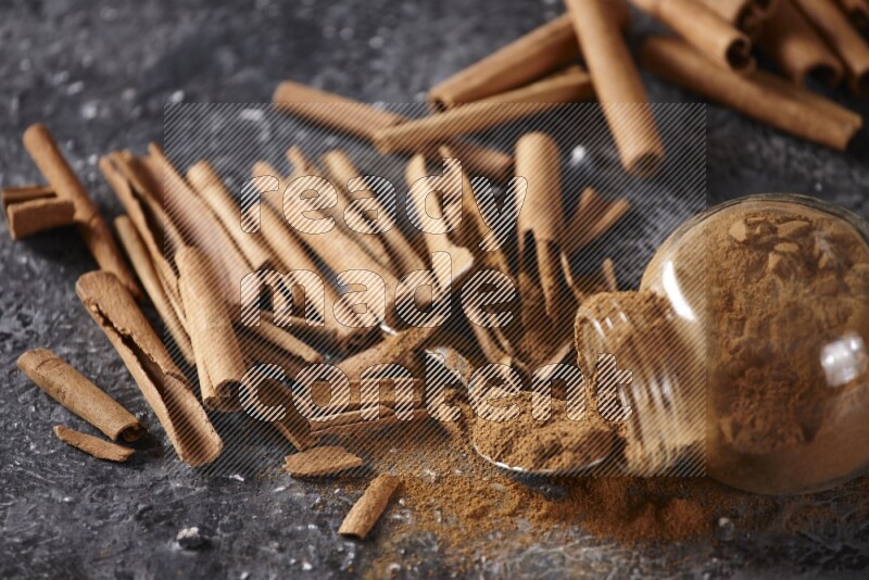 Herbal glass jar full cinnamon powder flipped and a metal spoon full of powder surrounded by cinnamon sticks on textured black background in different angles