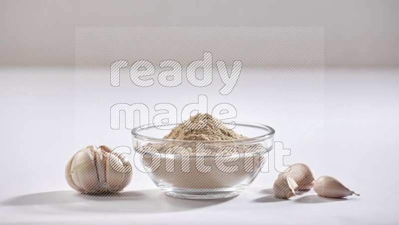 A glass bowl full of garlic powder with garlic bulb and some cloves beside it on a white flooring