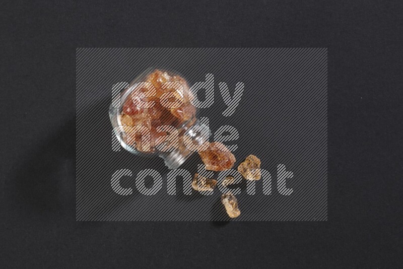 A glass spice jar full of gum arabic and jar is flipped with fallen pieces on black flooring