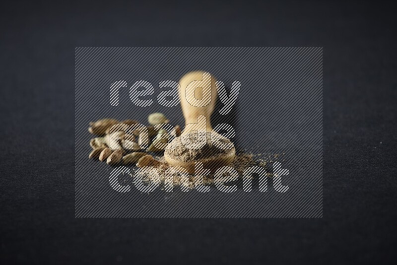 A wooden spoon full of cardamom powder and cardamom seeds beside it on black flooring