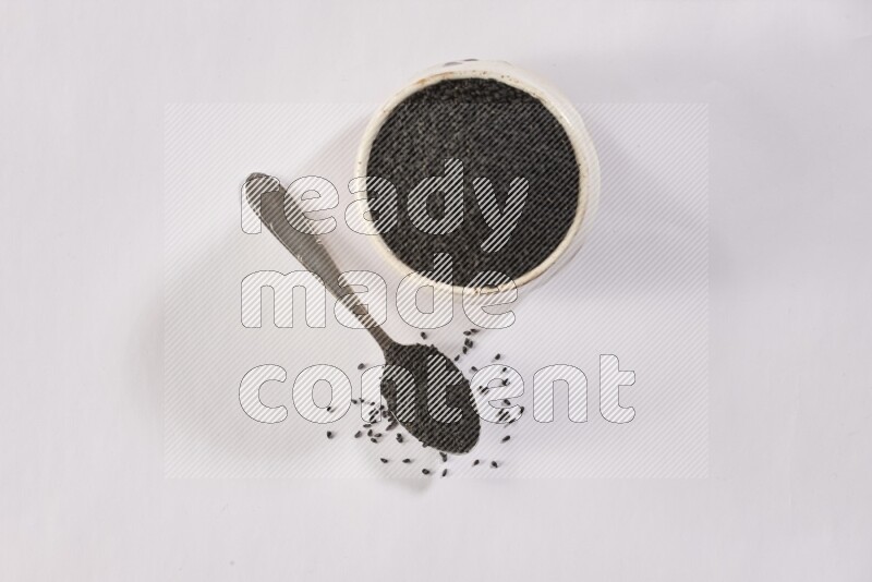 A beige pottery bowl and a metal spoon full of black seeds and more seeds spread on a white flooring