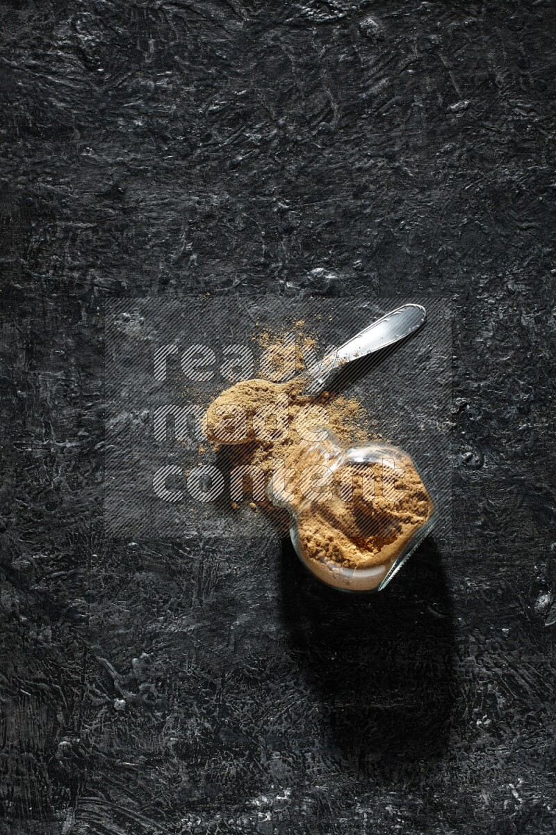 A flipped glass spice jar and a metal spoon full of allspice powder and powder spilled out of it on a textured black flooring