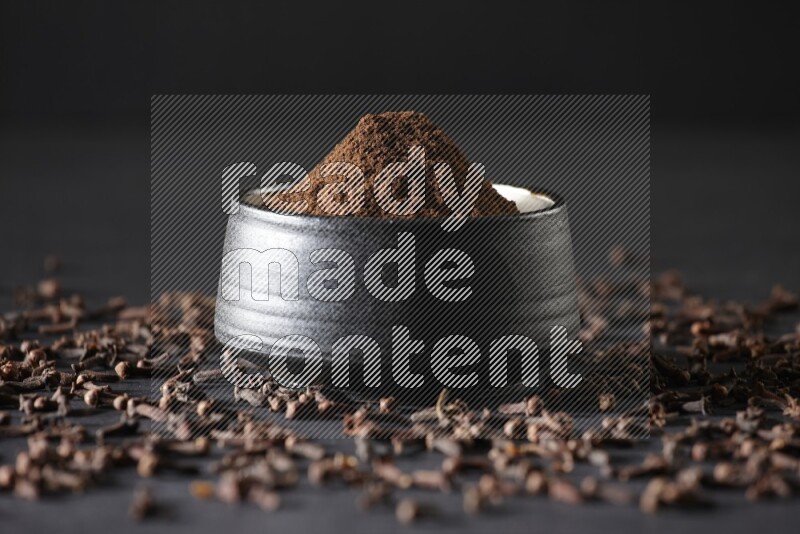 A black pottery bowl full of cloves powder and cloves whole grains on a black flooring