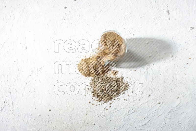 A flipped glass spice jar full of cumin powder and powder spilled out with cumin seeds on textured white flooring