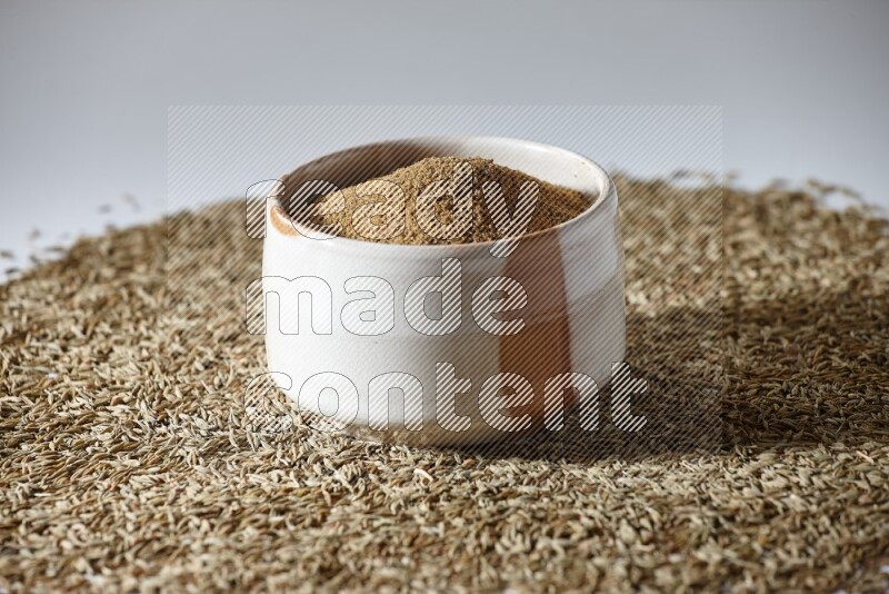 A beige bowl full of cumin powder with cumin seeds beneath it the bowl on a white flooring