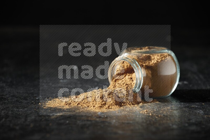 A flipped glass spice jar full of allspice powder and powder spilled out of it on a textured black flooring