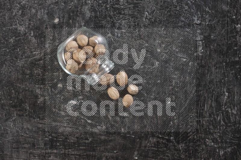 وعاء زجاجي ممتلئ بجوزة الطيب مقلوب وبعض الحبوب متناثرة على خلفية سوداء