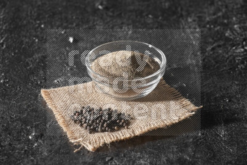 A glass bowl full of black pepper powder and black pepper beads on burlap fabric on textured black flooring