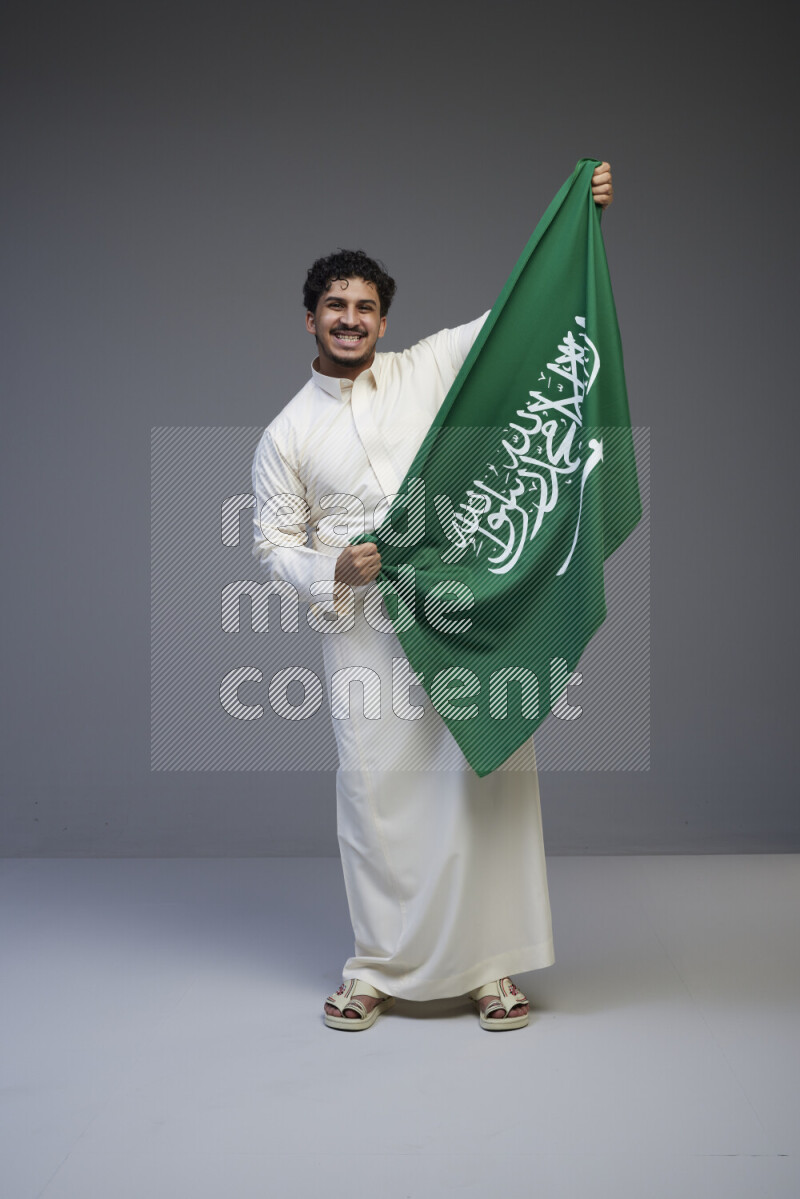 A Saudi man standing wearing thob holding big Saudi flag on gray background