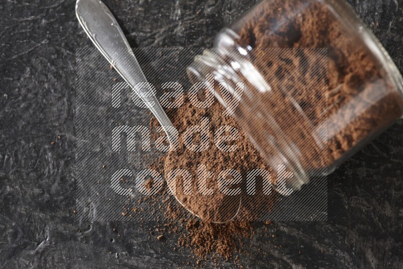 A flipped glass jar and metal spoon full of cloves powder with cloves spread on a textured black flooring