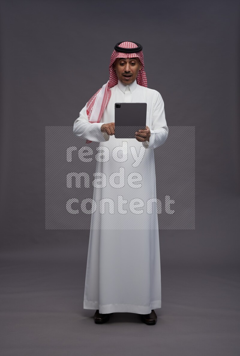 Saudi man wearing thob and shomag standing working on tablet on gray background