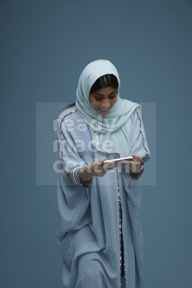 A Saudi woman Playing a Game on her phone on a blue background wearing a blue Abaya with hijab