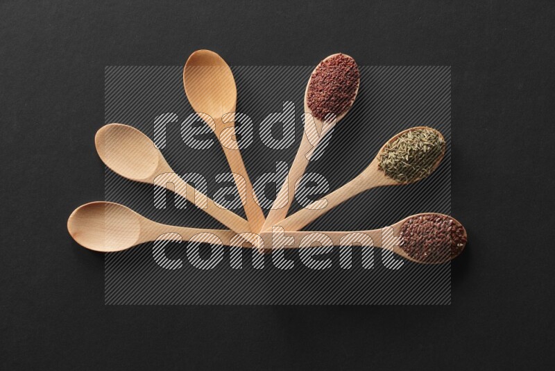 wooden spoons filled with white peppers, mustard seeds, black seeds, garden cress, cumin and flax on black flooring