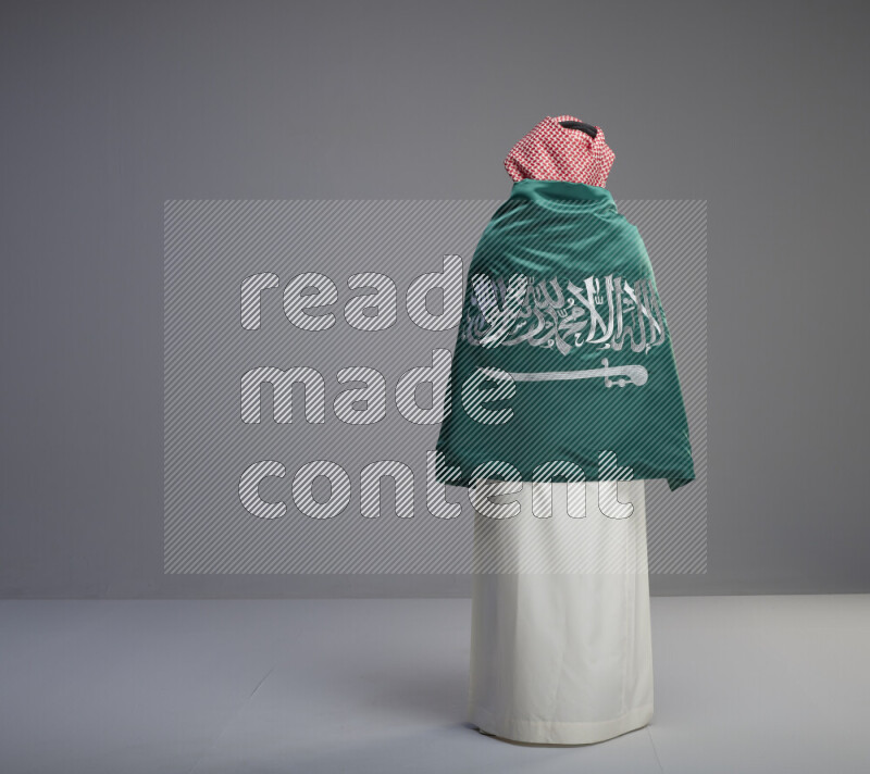 A Saudi man standing wearing thob and red shomag wrapping big saudi flag on gray background