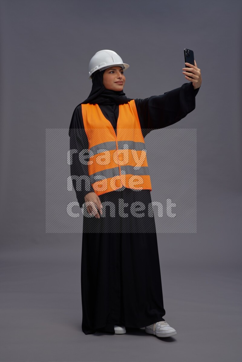 Saudi woman wearing Abaya with engineer vest standing taking selfie on gray background