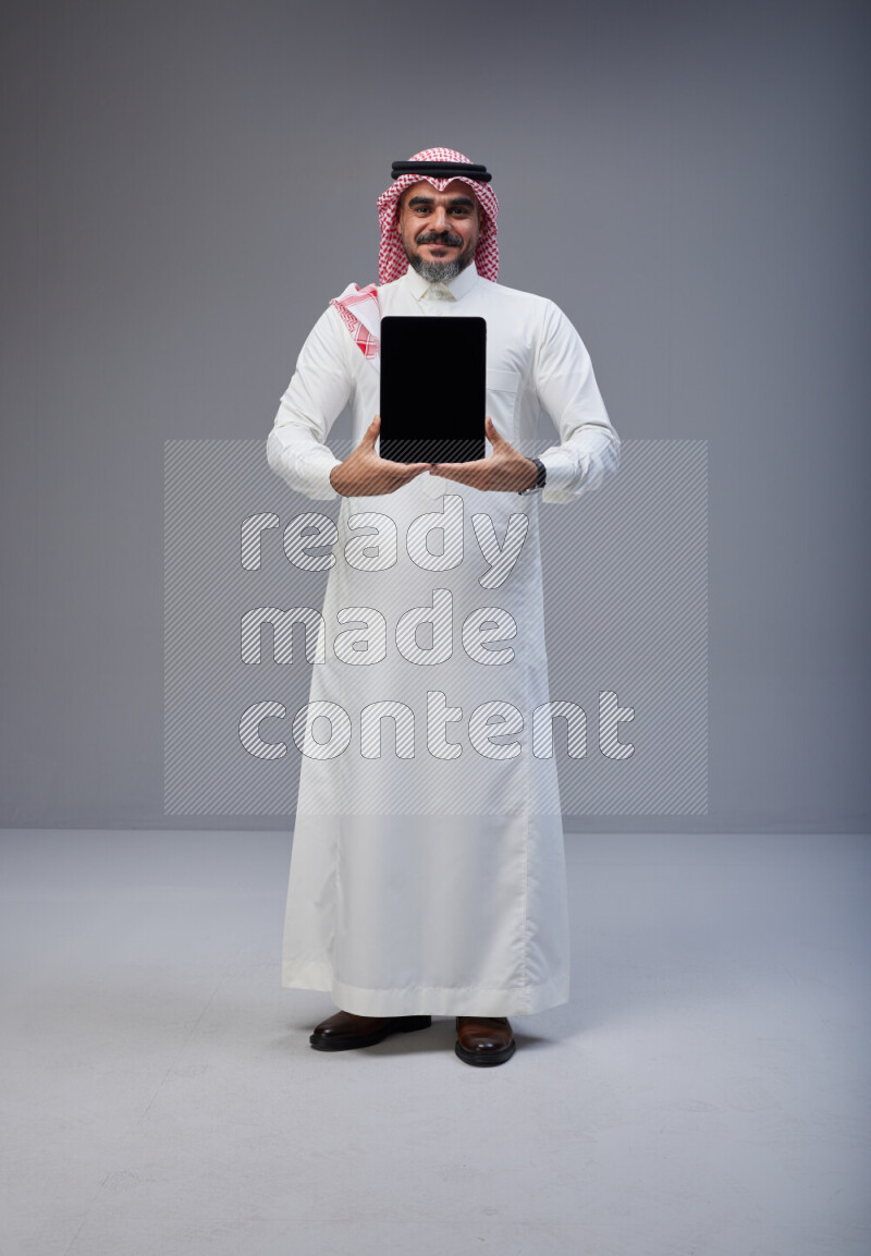 Saudi man Wearing Thob and red Shomag standing showing tablet to camera on Gray background