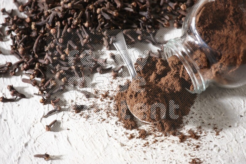 A flipped glass spice jar and a metal spoon full of cloves powder and powder came out of the jar with cloves spread on textured white flooring