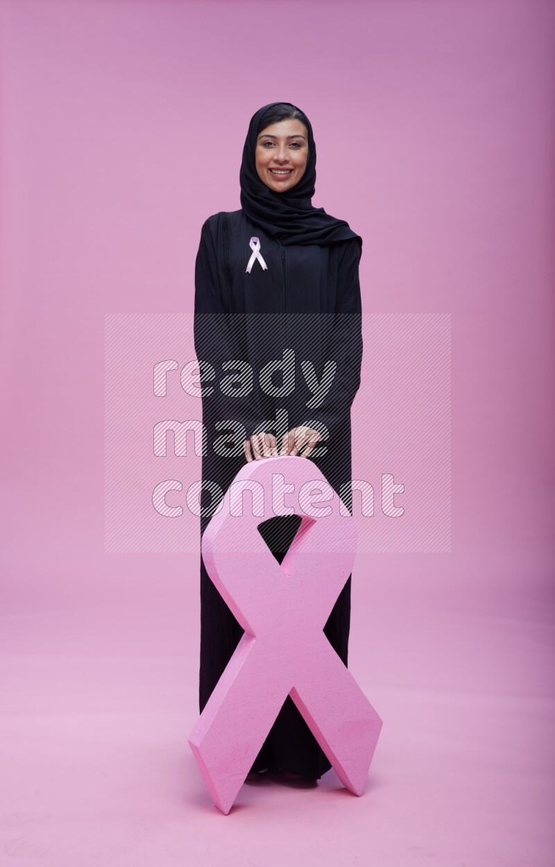 Saudi woman wearing pink ribbon on abaya standing holding awareness ribbon on plain pink background
