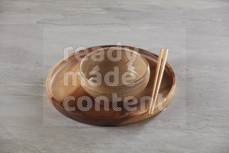 multi color pottery round bowl on top of brown wood round plate and wood chopsticks, on grey textured countertop
