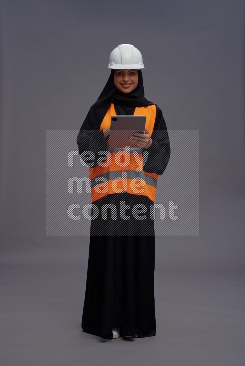 Saudi woman wearing Abaya with engineer vest standing working on tablet on gray background